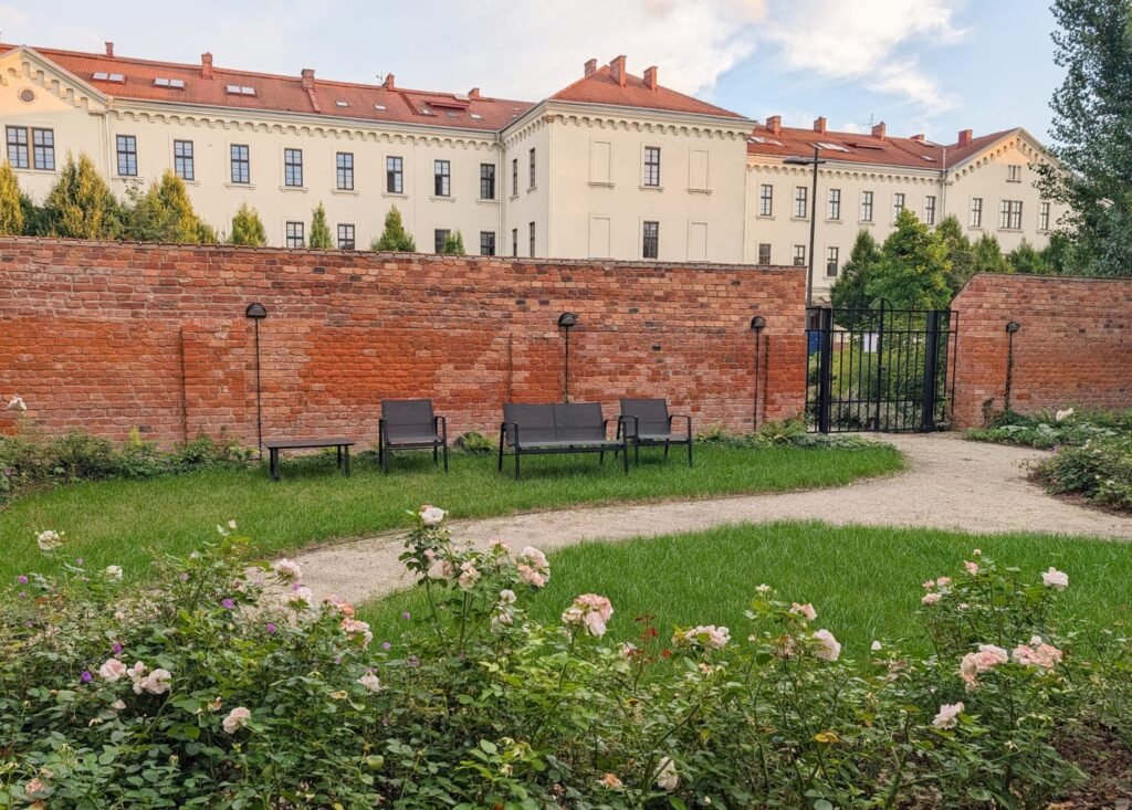 Ogród z zielenią, różami, alejką i meblami do siedzenia przy ceglanym murze, z historycznym budynkiem w tle, na eventy w Krakowie.